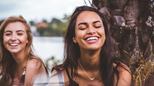 Two women happily laughing together outdoors by a serene lakeside.